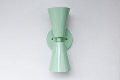 Mint Dryades Sconce in a fresh pastel green powder-coated finish, designed in the timeless midcentury modern double cone silhouette. This handcrafted steel wall light features two flared cones joined at a round backplate with warm brass accent screws, creating a sleek architectural form that casts light both upward and downward. The soft mint green color adds a soothing, playful touch to kitchens, bathrooms, hallways, or bedside walls, while the durable powder coat ensures lasting vibrancy. With its versati