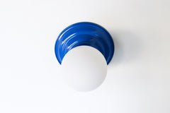 Bright Blue flushmount ceiling light or wall sconce with a frosted white globe.  Stepping back into a bygone era with a touch of modern minimalism, the Abeille Sconce or Flushmount is an artistic blend of form and function. This wall sconce doubles as a flushmount ceiling light, making it a versatile lighting solution for any space. Designed akin to a beehive, its charismatic shape is complemented by curated color options that add a playful element to its retro appeal.