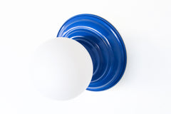 Bright Blue flushmount ceiling light or wall sconce with a frosted white globe.  Stepping back into a bygone era with a touch of modern minimalism, the Abeille Sconce or Flushmount is an artistic blend of form and function. This wall sconce doubles as a flushmount ceiling light, making it a versatile lighting solution for any space. Designed akin to a beehive, its charismatic shape is complemented by curated color options that add a playful element to its retro appeal.