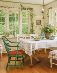 Bright farmhouse-style breakfast nook featuring the Adele chandelier with brass arms and sculptural shades, hanging above a rustic wood dining table covered in a blue-and-white checkered tablecloth. The cozy space includes green painted Windsor chairs with red cushions, floral arrangements, and surrounding windows framed with climbing ivy, blending modern handmade lighting with cottage-inspired dining design.