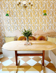 Vibrant kitchen breakfast nook featuring bold mustard-yellow botanical patterned wallpaper, mid-century modern brass sconces with adjustable green shades, and vintage-style brass chandelier. The space includes a wooden dining table, scalloped-edge built-in banquette seating with mustard velvet round pillows, striped green and yellow accent cushions, and a checkerboard floor in warm earthy tones, creating an inviting and eclectic dining area.