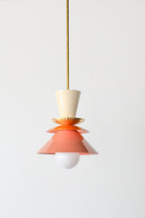 Close-up of the Astro Pendant by Sazerac Stitches, showcasing stacked powder-coated metal shades in cream, orange, and peach. A brass accent plate and white globe bulb complete this retro-inspired design.