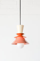 Close-up of the Astro Pendant by Sazerac Stitches, showcasing stacked powder-coated metal shades in cream, orange, and peach. A brass accent plate and white globe bulb complete this retro-inspired design.