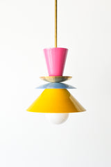 The Astro Pendant light by Sazerac Stitches, featuring a playful stacked design with bold enamel-coated metal shades in Doll Pink, Chrome or Brass, Hydrangea Blue, and Yellow. The geometric composition creates a striking visual contrast, suspended from a sleek rod with an exposed bulb for a modern touch.