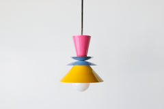 The Astro Pendant light by Sazerac Stitches, featuring a playful stacked design with bold enamel-coated metal shades in Doll Pink, Chrome or Brass, Hydrangea Blue, and Yellow. The geometric composition creates a striking visual contrast, suspended from a sleek rod with an exposed bulb for a modern touch.