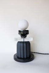Unique tabletop lamp viewed from an angle, showcasing the frosted glass globe, vintage reclaimed marble cup, thick clear acrylic disc, matte black ribbed ceramic body, and coordinating black cloth cord with inline switch; modern artistic lamp designed and handmade in New Orleans
