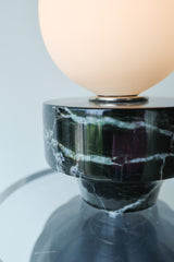 Sculptural table lamp glowing warmly with a frosted glass globe on top of a reclaimed black marble element, thick clear acrylic disc, and fluted matte black ceramic base; unique ambient lighting fixture handmade in New Orleans, perfect for a modern eclectic or artful interior
