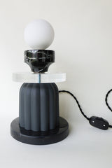 Unique tabletop lamp viewed from an angle, showcasing the frosted glass globe, vintage reclaimed marble cup, thick clear acrylic disc, matte black ribbed ceramic body, and coordinating black cloth cord with inline switch; modern artistic lamp designed and handmade in New Orleans