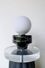 Unique tabletop lamp viewed from an angle, showcasing the frosted glass globe, vintage reclaimed marble cup, thick clear acrylic disc, matte black ribbed ceramic body, and coordinating black cloth cord with inline switch; modern artistic lamp designed and handmade in New Orleans