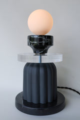 Sculptural table lamp glowing warmly with a frosted glass globe on top of a reclaimed black marble element, thick clear acrylic disc, and fluted matte black ceramic base; unique ambient lighting fixture handmade in New Orleans, perfect for a modern eclectic or artful interior