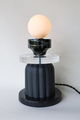 Handmade designer table lamp turned on, with a warm-glowing frosted globe atop layered materials including polished black marble, thick acrylic, and matte unglazed ceramic with vertical fluting; one-of-a-kind sculptural light for contemporary and maximalist interiors