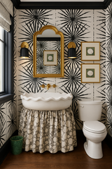 Eclectic powder room featuring a pair of matte black and brass wall sconces flanking a gold-framed mirror, mounted on bold black-and-white starburst wallpaper. The space includes a scalloped white sink with an upholstered skirt in a playful neutral polka dot pattern, a white toilet, warm wood flooring, and decorative wall art—styled to blend traditional elegance with graphic modern whimsy.