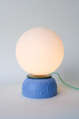 Modern bedside lamp with glowing white globe shade, matte blue ceramic base, olive rim, and twisted green cloth cord with inline switch. Small handmade artisan lamp ideal for nightstands, desks, or accent lighting in mid century modern, colorful, or eclectic home décor.