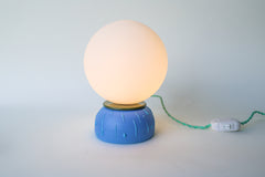 Modern bedside lamp with glowing white globe shade, matte blue ceramic base, olive rim, and twisted green cloth cord with inline switch. Small handmade artisan lamp ideal for nightstands, desks, or accent lighting in mid century modern, colorful, or eclectic home décor.