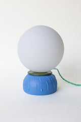 Unique ceramic globe table lamp featuring a matte blue base with textured drip design and olive green rim detail, paired with an opal white glass globe shade. Modern artisan lighting that works as a small bedside lamp, desk light, or accent lamp in contemporary, playful, or eclectic spaces.