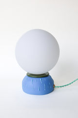 Unique ceramic globe table lamp featuring a matte blue base with textured drip design and olive green rim detail, paired with an opal white glass globe shade. Modern artisan lighting that works as a small bedside lamp, desk light, or accent lamp in contemporary, playful, or eclectic spaces.