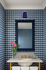 Bold powder room featuring cobalt blue Marcella wall sconce with fluted ceramic shade and brass hardware, mounted above a scalloped navy mirror. Blue gingham wallpaper wraps the space for a playful, preppy look. Marble sink with brass plumbing and pink flowers add contrast and charm.