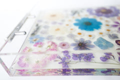 close up edge of tray Handmade acrylic tray with clear resin base and silver metal handles, featuring an intricate arrangement of real pressed flowers in soft pastels and rich jewel tones. The floral composition includes daisies, violets, cosmos, hydrangeas, and roses in a palette of purples, blues, lavenders, and blush pinks. The vibrant blossoms are carefully preserved in a dreamy, organic layout, creating a romantic, nature-inspired serving tray or decorative catchall perfect for spring, gift giving, or