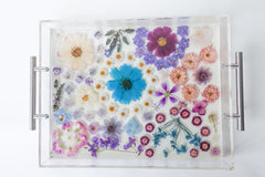 Handmade acrylic tray with clear resin base and silver metal handles, featuring an intricate arrangement of real pressed flowers in soft pastels and rich jewel tones. The floral composition includes daisies, violets, cosmos, hydrangeas, and roses in a palette of purples, blues, lavenders, and blush pinks. The vibrant blossoms are carefully preserved in a dreamy, organic layout, creating a romantic, nature-inspired serving tray or decorative catchall perfect for spring, gift giving, or adding a whimsical tou