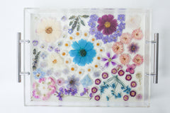 Handmade acrylic tray with clear resin base and silver metal handles, featuring an intricate arrangement of real pressed flowers in soft pastels and rich jewel tones. The floral composition includes daisies, violets, cosmos, hydrangeas, and roses in a palette of purples, blues, lavenders, and blush pinks. The vibrant blossoms are carefully preserved in a dreamy, organic layout, creating a romantic, nature-inspired serving tray or decorative catchall perfect for spring, gift giving, or adding a whimsical tou