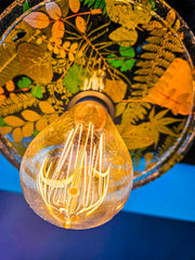 Close-up of a modern ceiling light fixture with three circular lampshades embedded with pressed leaves and greenery, creating a bright and artistic design against a white background.