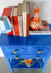 Bright blue nightstand with raw brass mushroom-shaped drawer pulls with orange books and a bedside Milton table lamp
