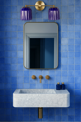 Bathroom vanity styled with the Double Marcella Sconce featuring two ribbed ceramic shades in a glossy cobalt blue glaze paired with a warm brass backplate and arms. Installed above a marble sink and gold-framed mirror against vibrant blue square tiles, this handmade wall light combines bold color, sculptural detail, and timeless craftsmanship. Designed by Sazerac Stitches in New Orleans, the double sconce is ideal for bathroom vanities, powder rooms, or modern eclectic interiors seeking colorful ceramic li
