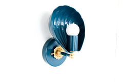 Shell-shaped wall sconce with a powdercoated lagoon blue finish and solid brass accents, including a detailed rosette and finial. The sconce features a sculptural shell form that cradles a bulb, casting soft ambient light. Shown from multiple angles on a white background, this coastal-inspired light fixture blends vintage Art Deco design with contemporary elegance, ideal for bedrooms, bathrooms, hallways, and eclectic interiors.