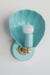 Shell-shaped wall sconce light fixture in a light aqua blue color with cast brass details.  Nancy Meyers aesthetic that also works with coastal or chinoiserie styled interiors.  Powdercoated steel with brass accents. Perfect wall sconce for powder rooms, bathrooms, and more.  Adds a little bit of fun whimsy with the nautical shell shape of the sconce.