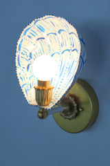 Close-up of hand-painted blue and white Coquille wall sconce with scalloped, shell-inspired shade made from powder-coated steel, softly illuminated with a warm bulb, mounted on a round brass backplate. The intricate blue brushstrokes and fluted edge details glow dramatically against a moody, dark background, highlighting the artisanal craftsmanship of this coastal-inspired, statement lighting fixture.