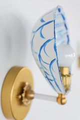 Close-up of hand-painted blue and white Coquille wall sconce with scalloped, shell-inspired shade made from powder-coated steel, softly illuminated with a warm bulb, mounted on a round brass backplate. The intricate blue brushstrokes and fluted edge details glow dramatically against a moody, dark background, highlighting the artisanal craftsmanship of this coastal-inspired, statement lighting fixture.