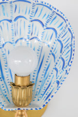 Angled view of a hand-painted powder-coated steel Coquille sconce featuring a white shell-shaped shade with vivid cobalt blue brushstroke accents, radiating from a central glowing bulb held in a vintage-style fluted brass socket. Mounted on a brushed brass wall plate with rosette detailing, the fixture pops against a soft blue wall, showcasing the perfect blend of coastal elegance and maximalist color in modern handmade lighting.