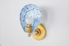 Side profile of a handmade wall sconce with a white powder-coated steel shade painted in bright blue brushstrokes, forming a scalloped shell silhouette. Mounted on an aged brass wall plate with vintage rosette detailing and a bold brass and ceramic socket arm, the sconce casts a soft glow, highlighting the custom-painted pattern and hand-finished brass elements. A playful yet elegant statement light for artful, color-forward spaces.