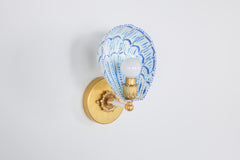 Side profile of a handmade wall sconce with a white powder-coated steel shade painted in bright blue brushstrokes, forming a scalloped shell silhouette. Mounted on an aged brass wall plate with vintage rosette detailing and a bold brass and ceramic socket arm, the sconce casts a soft glow, highlighting the custom-painted pattern and hand-finished brass elements. A playful yet elegant statement light for artful, color-forward spaces.