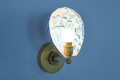 Side profile of a handmade wall sconce with a white powder-coated steel shade painted in bright blue brushstrokes, forming a scalloped shell silhouette. Mounted on an aged brass wall plate with vintage rosette detailing and a bold brass and ceramic socket arm, the sconce casts a soft glow, highlighting the custom-painted pattern and hand-finished brass elements. A playful yet elegant statement light for artful, color-forward spaces.