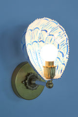 Side profile of a handmade wall sconce with a white powder-coated steel shade painted in bright blue brushstrokes, forming a scalloped shell silhouette. Mounted on an aged brass wall plate with vintage rosette detailing and a bold brass and ceramic socket arm, the sconce casts a soft glow, highlighting the custom-painted pattern and hand-finished brass elements. A playful yet elegant statement light for artful, color-forward spaces.