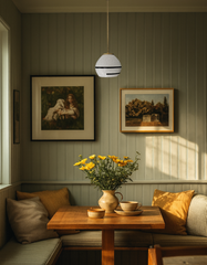 Warm and inviting breakfast nook featuring the handmade Delta pendant light in white ceramic with black accents, hanging above a wooden dining table with striped banquette seating and golden throw pillows. The corner dining space includes vintage-style wall art, natural light, and a rustic vase of yellow flowers, blending artisanal lighting with cozy modern cottage design.