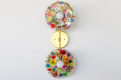 Modern two light wall sconce with brass hardware. Features resin-encapsulated multi colored flowers & greenery, ferns, and leaves.  Adds a real-like nature theme to any interior design project.  Called the Botanico Sconce by Sazerac Stitches. Features flowers in every colors, green & yellow leaves.