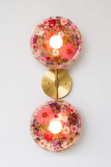 Modern two light wall sconce with brass hardware and pink & purple flowers. Features resin-encapsulated pink & purple flowers with greenery, ferns, and leaves.  Adds a real-like nature theme to any interior design project.  Called the Botanico Sconce by Sazerac Stitches. Features pink & purple flowers with greenery