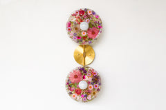 Modern two light wall sconce with brass hardware and pink & purple flowers. Features resin-encapsulated pink & purple flowers with greenery, ferns, and leaves.  Adds a real-like nature theme to any interior design project.  Called the Botanico Sconce by Sazerac Stitches. Features pink & purple flowers with greenery