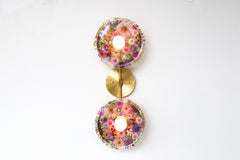 Modern two light wall sconce with brass hardware and pink & purple flowers. Features resin-encapsulated pink & purple flowers with greenery, ferns, and leaves.  Adds a real-like nature theme to any interior design project.  Called the Botanico Sconce by Sazerac Stitches. Features pink & purple flowers with greenery