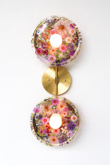 Modern two light wall sconce with brass hardware and pink & purple flowers. Features resin-encapsulated pink & purple flowers with greenery, ferns, and leaves.  Adds a real-like nature theme to any interior design project.  Called the Botanico Sconce by Sazerac Stitches. Features pink & purple flowers with greenery