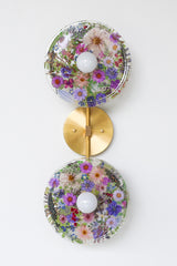 Modern two light wall sconce with brass hardware and purple flowers. Features resin-encapsulated purple flowers with greenery, ferns, and leaves.  Adds a real-like nature theme to any interior design project.  Called the Botanico Sconce by Sazerac Stitches. Features purple flowers with greenery