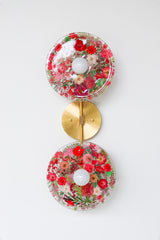 Modern two light wall sconce with brass hardware and red flowers. Features resin-encapsulated pink & purple flowers with greenery, ferns, and leaves.  Adds a real-like nature theme to any interior design project.  Called the Botanico Sconce by Sazerac Stitches. Features red flowers with greenery