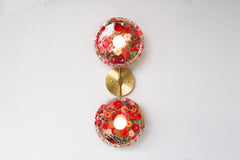 Modern two light wall sconce with brass hardware and red flowers. Features resin-encapsulated pink & purple flowers with greenery, ferns, and leaves.  Adds a real-like nature theme to any interior design project.  Called the Botanico Sconce by Sazerac Stitches. Features red flowers with greenery