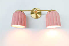 Double Marcella Sconce by Sazerac Stitches featuring two pastel pink ceramic shades with scalloped ridges, glowing warmly against a brass backplate. Handmade in the USA, this feminine Art Deco-inspired fixture adds charm and color to bathrooms, vanities, and bedrooms while providing compact yet stylish lighting.