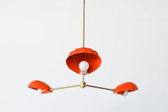 The Coquille Chandelier in Flame is a bold, sculptural light fixture featuring three scallop shell-shaped shades in a vibrant red-orange powder-coated finish. Suspended from a central brass rod, each shell houses a globe bulb and radiates from a sleek brass frame, combining coastal lighting style with vintage Art Deco influence. This modern chandelier is handcrafted for statement interiors and is perfect for dining rooms, bedrooms, or eclectic spaces seeking designer lighting with color and personality.