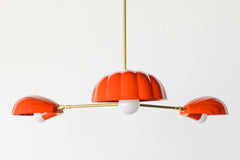 The Coquille Chandelier in Flame is a bold, sculptural light fixture featuring three scallop shell-shaped shades in a vibrant red-orange powder-coated finish. Suspended from a central brass rod, each shell houses a globe bulb and radiates from a sleek brass frame, combining coastal lighting style with vintage Art Deco influence. This modern chandelier is handcrafted for statement interiors and is perfect for dining rooms, bedrooms, or eclectic spaces seeking designer lighting with color and personality.