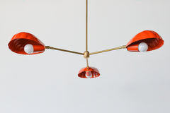 The Coquille Chandelier in Flame is a bold, sculptural light fixture featuring three scallop shell-shaped shades in a vibrant red-orange powder-coated finish. Suspended from a central brass rod, each shell houses a globe bulb and radiates from a sleek brass frame, combining coastal lighting style with vintage Art Deco influence. This modern chandelier is handcrafted for statement interiors and is perfect for dining rooms, bedrooms, or eclectic spaces seeking designer lighting with color and personality.