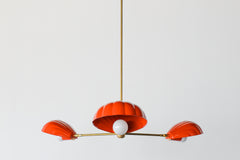 The Coquille Chandelier in Flame is a bold, sculptural light fixture featuring three scallop shell-shaped shades in a vibrant red-orange powder-coated finish. Suspended from a central brass rod, each shell houses a globe bulb and radiates from a sleek brass frame, combining coastal lighting style with vintage Art Deco influence. This modern chandelier is handcrafted for statement interiors and is perfect for dining rooms, bedrooms, or eclectic spaces seeking designer lighting with color and personality.